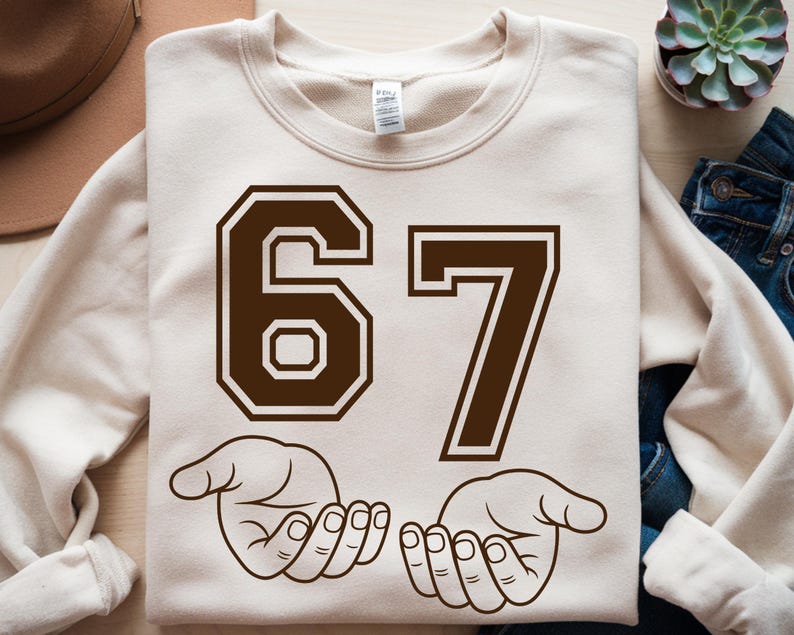 Pode incluir: Camisola creme com o n&uacute;mero 67 em castanho, com uma fonte de estilo universit&aacute;rio. Abaixo dos n&uacute;meros, duas m&atilde;os abertas em contorno castanho. Uma pe&ccedil;a de roupa casual e confort&aacute;vel.