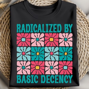 Op de afbeelding: Zwart T-shirt met de tekst "RADICALIZED BY BASIC DECENCY" in turquoise. Het shirt heeft een raster van kleurrijke bloemmotieven in roze, turquoise en geel. Het shirt wordt gepresenteerd op een geweven oppervlak.