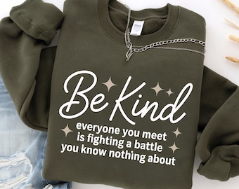 Könnte beinhalten: Ein olivgrünes Sweatshirt mit dem weißen Schriftzug "Be Kind" und dem Text "everyone you meet is fighting a battle you know nothing about". Eine silberne Kette liegt über der Schulter.