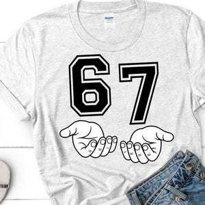 K&ouml;nnte beinhalten: Ein hellgraues T-Shirt mit der Zahl "67" in fetter, schwarzer Schrift im College-Stil. Darunter befinden sich zwei offene H&auml;nde in einer Strichzeichnung. Das T-Shirt besteht aus einem weichen Material.