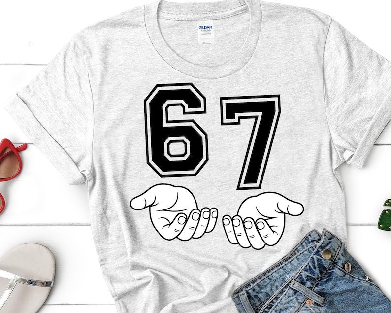 K&ouml;nnte beinhalten: Ein hellgraues T-Shirt mit der Zahl "67" in fetter, schwarzer Schrift im College-Stil. Darunter befinden sich zwei offene H&auml;nde in einer Strichzeichnung. Das T-Shirt besteht aus einem weichen Material.