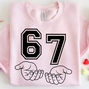 Pode incluir: Camisola rosa claro com o n&uacute;mero 67 em preto, contornado a branco. Abaixo dos n&uacute;meros, duas m&atilde;os pretas com as palmas abertas. A camisola est&aacute; dobrada, com uma etiqueta vis&iacute;vel no interior.