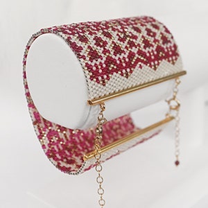 May include: A beaded cuff bracelet in red, white, and gold, displayed on a white cylindrical form. The bracelet features a geometric pattern and gold-coloured metal accents with a chain closure. The design is intricate and detailed.