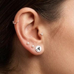 Könnte beinhalten: Ein Ohr ist mit mehreren silbernen Ohrringen geschmückt. Dazu gehören ein kleiner Reifen, ein Schmetterlingsstecker, zwei kleine Perlenstecker und ein runder Stecker mit dem schwarzen Buchstaben "A". Das Ohr befindet sich vor einem neutralen Hintergrund.