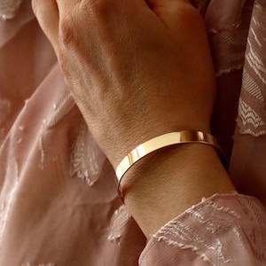 Pode incluir: Um bracelete de ouro simples com um acabamento liso e polido. O bracelete é aberto e ajustável para se ajustar à maioria dos pulsos.