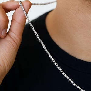 Op de afbeelding: Een zilveren ketting met een afgerond box chain ontwerp. De ketting wordt door een hand tegen een zwarte achtergrond gehouden, waarbij de reflecterende kwaliteit en de ingewikkelde schakels van de ketting worden benadrukt. Een klassiek sieraad.