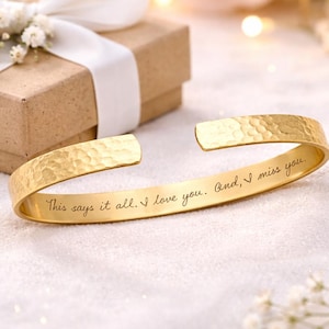 Peut inclure: Un bracelet manchette doré avec une texture martelée. Le bracelet a un design ouvert et porte le message gravé : "This says it all. I love you. And, I miss you." Un petit coffret cadeau avec un ruban blanc est en arrière-plan.