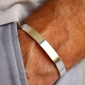 Könnte beinhalten: Ein poliertes silbernes Armband. Das Armband hat ein flaches, rechteckiges Design und wird am Handgelenk getragen. Das Armband ist etwa 1,3 cm breit und hat eine reflektierende Oberfläche.
