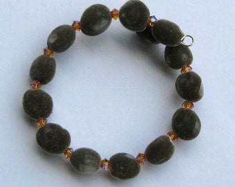 May include: A bracelet featuring olive-green, oval-shaped beads interspersed with small, orange-toned crystals. The bracelet is strung on a flexible wire and has a silver-colored clasp. The beads have a velvety texture.