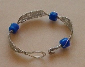 May include: A silver wire bracelet with three bright blue square beads. The bracelet is open, showing the clasp. The wire is woven in a delicate pattern.