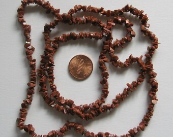 May include: A long strand of brown and gold sunstone chip beads. The beads are small and irregular in shape. The strand is laid out in a circle, with a penny in the centre for scale.