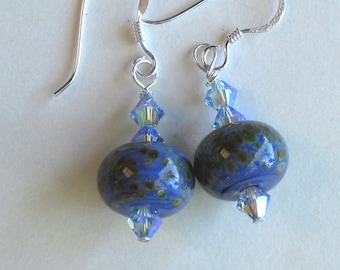 May include: A pair of blue glass bead earrings with silver hooks. The beads feature a speckled pattern of brown and green. Clear, faceted crystals are placed above and below the blue beads. The earrings are displayed on a white surface.