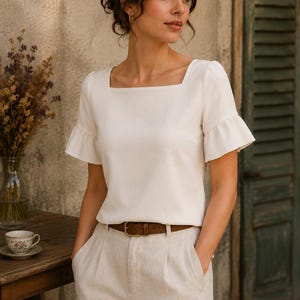 Puede incluir: Una blusa de color crema de manga corta con escote cuadrado y bordes de manga con volantes, combinada con pantalones de color beige claro y un cinturón de cuero marrón con hebilla dorada, creando un conjunto de tonos neutros y líneas limpias.