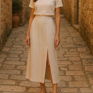 Può includere: Una donna indossa una blusa e una gonna in lino beige con uno spacco sul davanti. La gonna è al ginocchio e la blusa ha maniche corte. È in piedi in un vicolo di pietra.