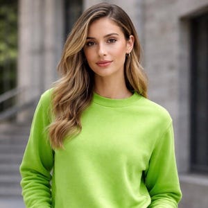 Könnte beinhalten: Ein limettengrünes Sweatshirt mit Rundhalsausschnitt und langen Ärmeln. Das Sweatshirt wird mit hellblauen Jeans kombiniert. Das Model hat lange, wellige blonde Haare und steht draußen mit den Händen in den Taschen.