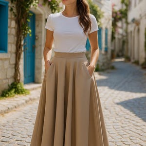 Könnte beinhalten: Eine Frau trägt ein weißes T-Shirt, eine Sonnenbrille und einen langen, beigefarbenen Rock mit Taschen. Der Rock reicht bis zu den Knöcheln. Im Hintergrund eine Kopfsteinpflasterstraße und Gebäude mit blauen Fensterläden.