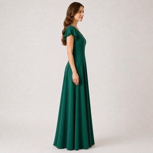 May include: A long, emerald green dress with a V-neckline and short, ruffled sleeves. The dress has a flowing A-line skirt. The model is standing in profile against a neutral background.