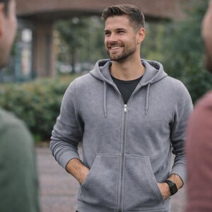 Peut inclure: Un sweat à capuche gris clair zippé avec un cordon de serrage, des manches longues et deux poches avant. Le sweat est porté sur un t-shirt gris foncé. Une montre numérique noire est visible au poignet. Le vêtement a un design simple et fonctionnel.