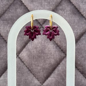Peut inclure: Une paire de boucles d'oreilles créoles avec de petites breloques en forme de feuille violet foncé. Les boucles sont composées de fins anneaux en métal doré poli retenant les pendentifs texturés en forme de feuille, aux bords dentelés.