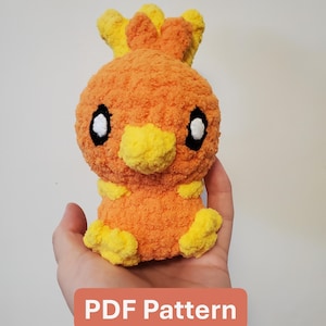 May include: A plush orange and yellow Torchic character, with black and white eyes, is held in a hand. The plushie has a textured, fuzzy appearance. The text "PDF Pattern" is displayed on a rectangular orange background.