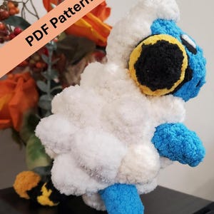 May include: A plush, handmade sheep-like creature with white, blue, yellow, and black accents. The fluffy toy has a round body and is labeled "PDF Pattern" in a diagonal banner. The toy is likely a crochet or knit project.
