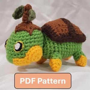 May include: A crocheted turtle figurine in shades of green, brown, and yellow. The turtle has a detailed shell and a leaf-like feature on its head. The text "PDF Pattern" is displayed on a rectangular orange background.