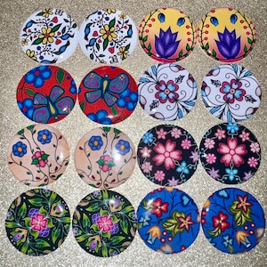 May include: A collection of sixteen round, decorative buttons with various floral and butterfly designs. The buttons feature vibrant colors like red, blue, pink, and green, with intricate patterns. The buttons are arranged on a gold glitter surface.