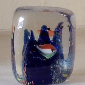 May include: A clear glass paperweight with a blue, orange, and white glass sculpture of fish inside. The sculpture is surrounded by clear glass.