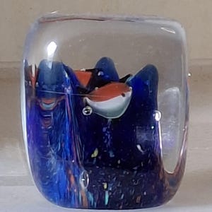 May include: A glass paperweight with a blue, orange, and white abstract design inside. The paperweight is clear and has a square shape.