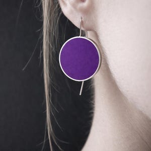 Könnte beinhalten: Nahaufnahme eines silbernen Ohrrings mit einem kreisförmigen, tiefvioletten Mittelteil. Der Ohrring hat ein schlichtes, modernes Design mit einem dünnen Silberrahmen und einem geraden Silberstift. Der Ohrring hängt an einem Ohr.