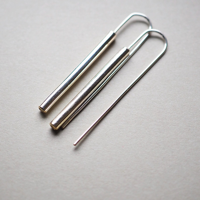 Puede incluir: Un par de pendientes de aro de plata con un dise&ntilde;o largo, delgado y recto. Los pendientes est&aacute;n hechos de metal y tienen un estilo simple y minimalista.