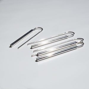 Puede incluir: Un conjunto de cinco pendientes de plata, cada uno con una forma cil&iacute;ndrica larga y delgada y un bucle en un extremo. Los pendientes est&aacute;n dispuestos en un patr&oacute;n escalonado sobre una superficie blanca.