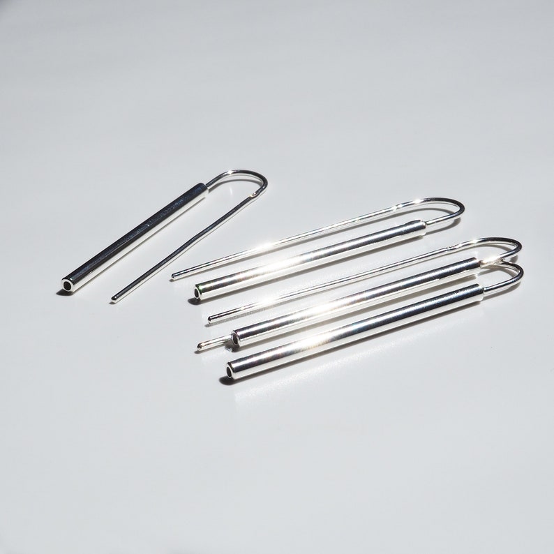 Puede incluir: Un conjunto de cinco pendientes de plata, cada uno con una forma cil&iacute;ndrica larga y delgada y un bucle en un extremo. Los pendientes est&aacute;n dispuestos en un patr&oacute;n escalonado sobre una superficie blanca.