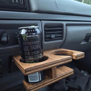 May include: A wooden cup holder with two circular cutouts attached to a vehicle dashboard. A black Monster Energy drink can is placed in one of the circular openings. The holder features a layered wood construction with a smooth, stained finish.