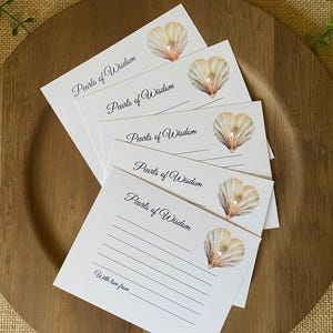 May include: A stack of white cards with the text "Pearls of Wisdom" and a seashell illustration with a pearl. The cards are arranged on a wooden plate, with a burlap background and greenery.