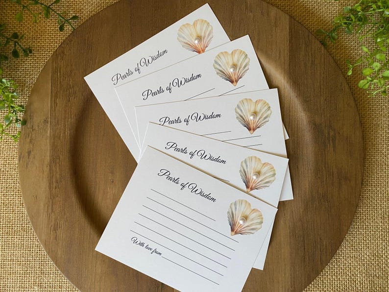 May include: A stack of white cards with the text "Pearls of Wisdom" and a seashell illustration with a pearl. The cards are arranged on a wooden plate, with a burlap background and greenery.