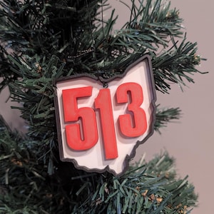May include: A Christmas tree ornament in the shape of Ohio with the area code 513 in red. The ornament has a white background and a black outline, hanging from a black string. The ornament is hanging on a green Christmas tree.
