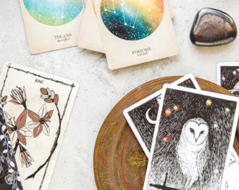 May include: A set of tarot cards with illustrations of a king, an owl, and constellations. The cards are laid out on a white surface with a small black stone and a decorative plate.