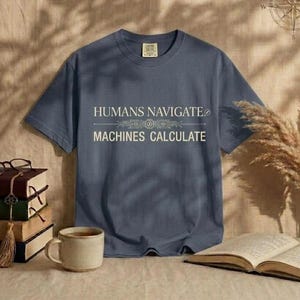 Peut inclure: Un t-shirt bleu ardoise à manches courtes avec le texte blanc "HUMANS NAVIGATE" au-dessus d'un graphique de circuit imprimé horizontal et le texte "MACHINES CALCULATE" en dessous. Le t-shirt a un col rond et une étiquette de marque rectangulaire.