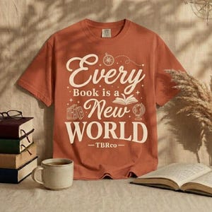 Peut inclure: Un t-shirt à manches courtes de couleur rouille avec le texte crème Every Book is a New World, accompagné d'illustrations d'une boussole, d'un livre ouvert, d'une pile de livres et d'un globe, avec le petit texte TBRco imprimé en bas.