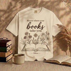 Peut inclure: Un t-shirt à manches courtes couleur crème avec un motif marron foncé représentant un livre ouvert d'où poussent des fleurs. Le texte "Bloom where your books take you" est inscrit au-dessus et "TBRco" est centré sous le livre.