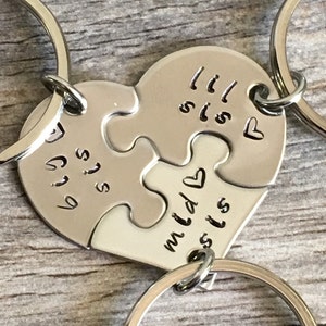 May include: Two silver keychains with heart-shaped puzzle pieces that say "Big Sis" and "Lil Sis" on them. The keychains are connected by a silver ring.
