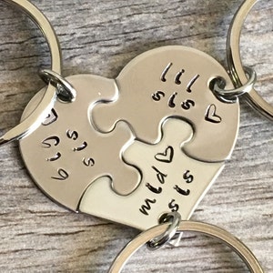 May include: Two silver keychains with heart-shaped puzzle pieces that say "Big Sis" and "Lil Sis" on them. The keychains are connected by a silver ring.