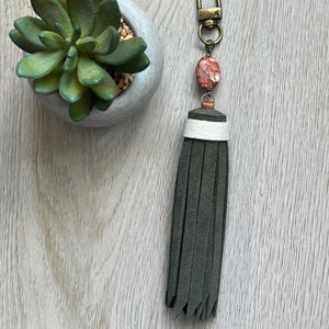 May include: A green suede tassel keychain with a white and brown accent and a red and white stone charm. The keychain is attached to a gold metal key ring.