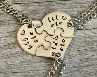 May include: Three silver necklaces with heart-shaped puzzle pieces that say "lil sis", "mid sis", and "big sis" on them.