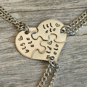 May include: Three silver necklaces with heart-shaped puzzle pieces that say "lil sis", "mid sis", and "big sis" on them.