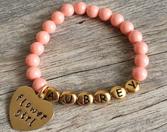 May include: A pink beaded bracelet with a gold heart charm that says "flower girl" and gold letter beads that spell "AUBREY".