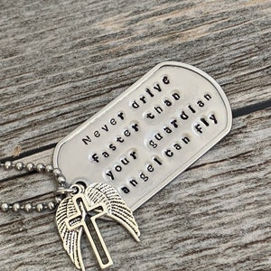 May include: Silver necklace with a dog tag pendant that reads "Never drive faster than your guardian angel can fly". The dog tag is attached to a silver chain with a silver charm of a cross with wings.