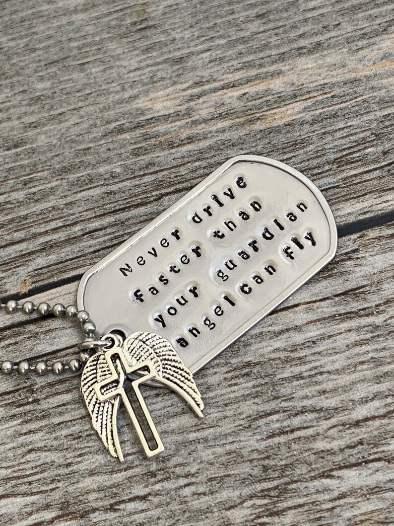 May include: Silver necklace with a dog tag pendant that reads "Never drive faster than your guardian angel can fly". The dog tag is attached to a silver chain with a silver charm of a cross with wings.