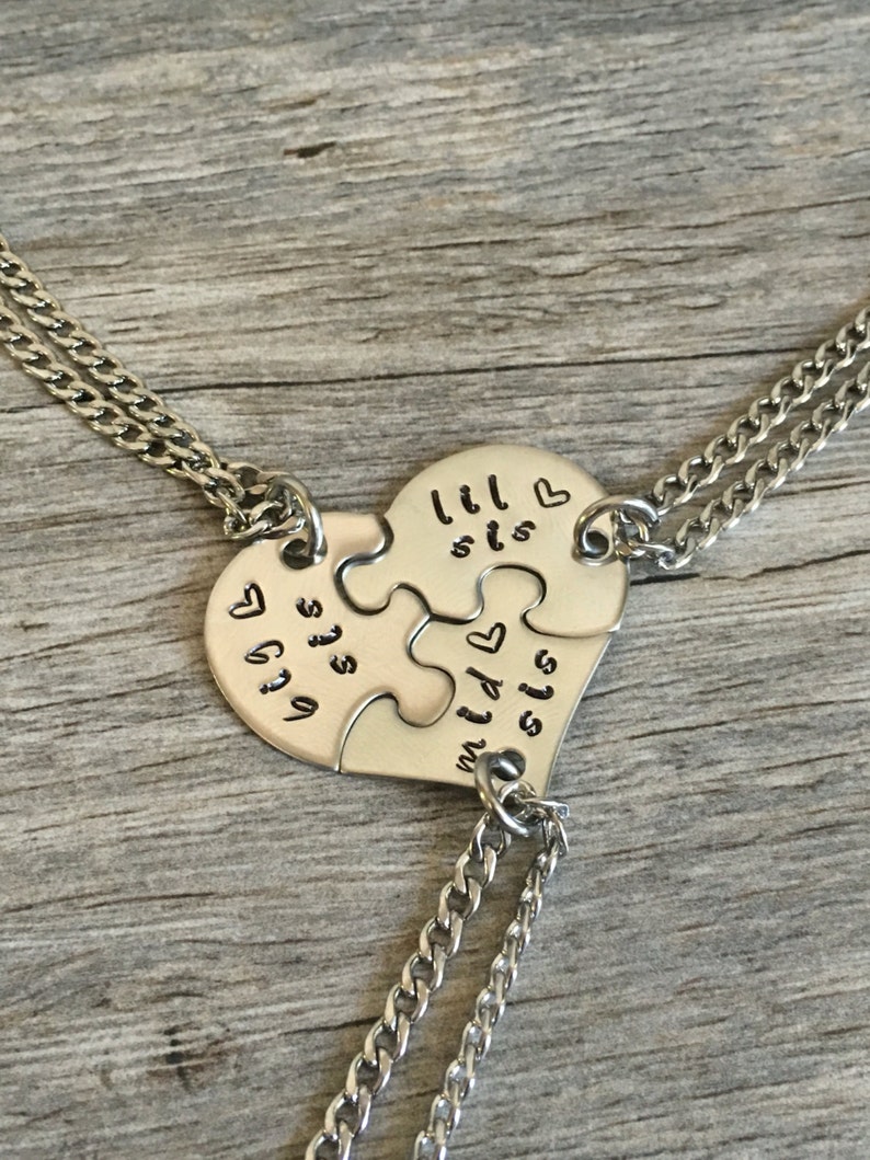 May include: Three silver necklaces with heart-shaped puzzle pieces that say "lil sis", "mid sis", and "big sis" on them.